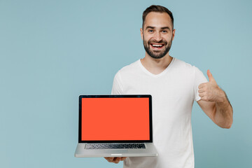 Young smiling man in white blank print design t-shirt hold use work on laptop pc computer with blank screen workspace area show thumb up gesture isolated on plain pastel light blue background studio.