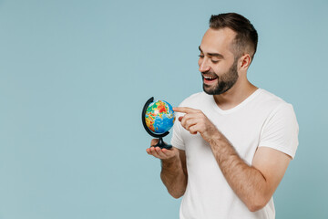 Young smiling happy geography teacher man 20s wear casual white t-shirt hold in hands touch hoose country spin Earth world globe isolated on plain pastel light blue color background studio portrait