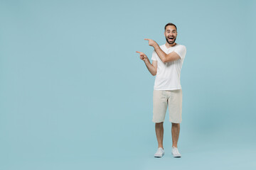 Fototapeta na wymiar Full length young satisfied overjoyed happy man 20s wear casual white t-shirt point index finger aside on workspace area mock up isolated on plain pastel light blue color background studio portrait