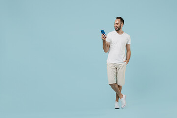 Fototapeta na wymiar Full length young smiling friendly happy man 20s wearing casual white t-shirt looking camera using mobile cell phone chat onine isolated on plain pastel light blue color background studio portrait