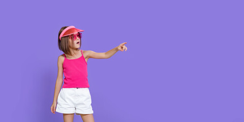 Little girl in white shorts, a pink T-shirt, pink trendy glasses and a summer visor pointing finger to empty space on a purple background in the studio.