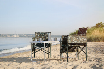 Camouflage fishing chairs and table with metal cups on sandy beach near river