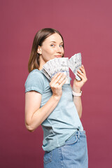 Funny young lady in a blue summer tshirt is holding money and looking to them over a pink background with copy space