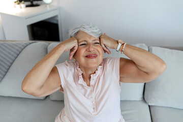 Photo of a senior woman suffering from headache while sitting on couch in a living room. Depressed mature woman with head in hand feeling strong pain. Stressed old lady suffering from migraine at home