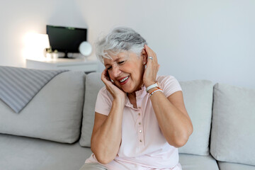 Photo of a senior woman suffering from headache while sitting on couch in a living room. Depressed mature woman with head in hand feeling strong pain. Stressed old lady suffering from migraine at home