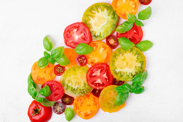Slices of assorted tomatoes of different colors and sizes and basil leaves on white table. Flat lay. Food background