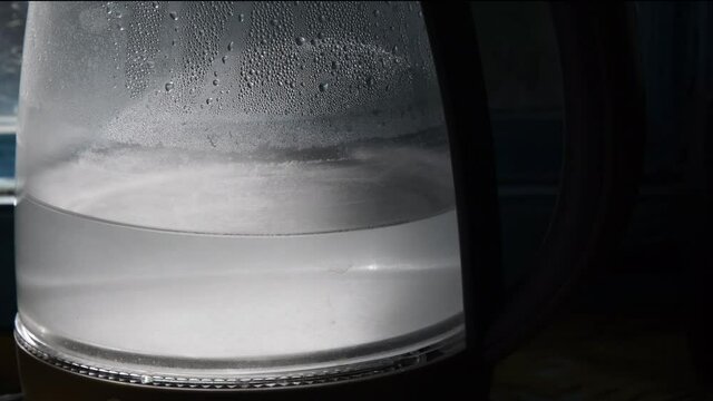 Sunlight shines in hot water inside teapot with blurry condensation on glass surface. Turbid water waving inside old transparent kettle in kitchen