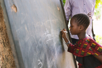 Concentrated little black girl writing numbers on a blackboard during open air classes in a rural...