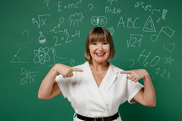 Confident teacher mature elderly senior lady woman 55 wear white shirt point index fingers on herself isolated green wall chalk blackboard background studio. Education in school September 1 concept