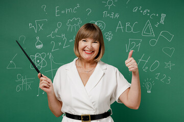 Excited smiling fun teacher mature elderly lady woman 55 wear white shirt hold in hand demonstrate with pointer show thumb up like gesture isolated on green wall chalk blackboard background studio