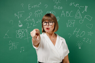 Angry teacher elderly lady woman 55 wear shirt glasses point index finger camera on you motivating encourage isolated on green wall chalk blackboard background studio. Education high school concept