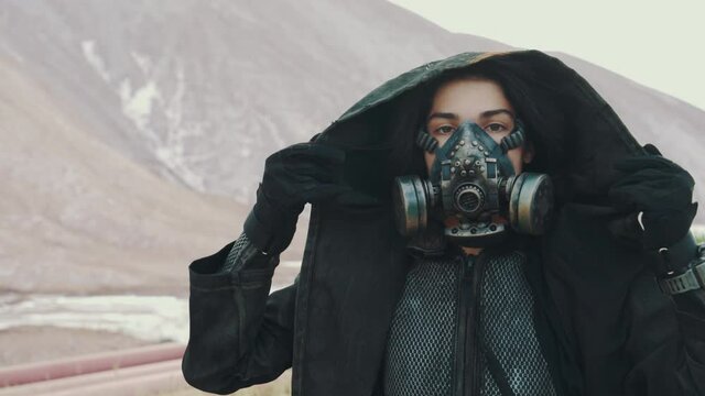 Portrait post apocalyptic woman in grunge clothes hooded mantle and protective gas mask posing by white hills in desert looking into camera. Future female survivor. Post apocalyptic world concept