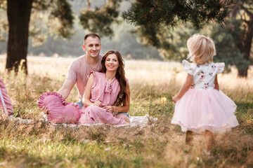 Naklejka premium Mom, dad and little daughter are sitting next to wigwam decoration in the park. Family spending time outdoor in summer, having fun together. Girl are dressed in pink dress