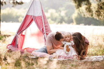 Mom, dad and little daughter are sitting next to wigwam decoration in the park. Family spending time outdoor in summer, having fun together. Girl are dressed in pink dress © Andriy Medvediuk