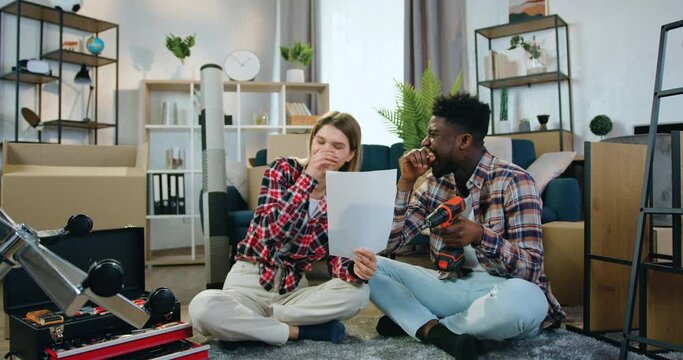 Attractive smiling modern young multiracial couple sitting on the floor in new flat and planning room design using ready scheme on sheet of paper