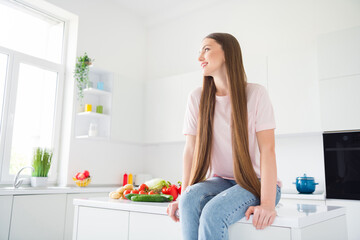 Portrait of attractive cheery dreamy long-haired girl sitting on countertop cooking in light white home kitchen indoors