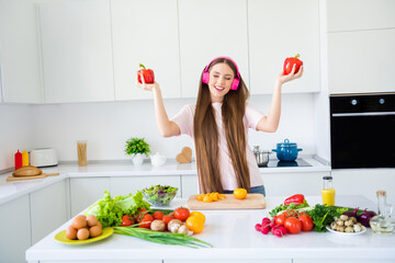 Portrait of attractive cheery dreamy girl cooking yummy omestic salad listening sound dancing in light white home kitchen indoors