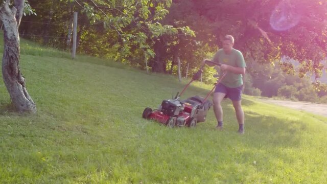 Man mowing lawn between fruit trees with red petrol rotary lawnmower machine. Aerial 4k view.
