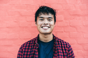 Happy Asian guy portrait - Confident asian young male having fun smiling while posing in front of camera against a red wall background - obrazy, fototapety, plakaty