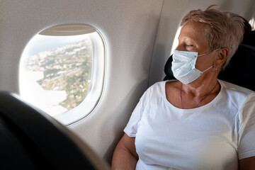 An elderly woman travels. Flying in a plane wearing a mask. Passenger. Watch the porthole. Look at the ground from above. Active lifestyle.