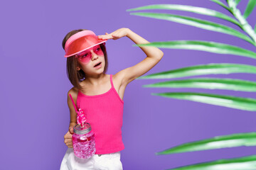 a little girl in white shorts, a pink T-shirt, pink trendy glasses, with a drink in hand and a summer visor on a purple background in the studio