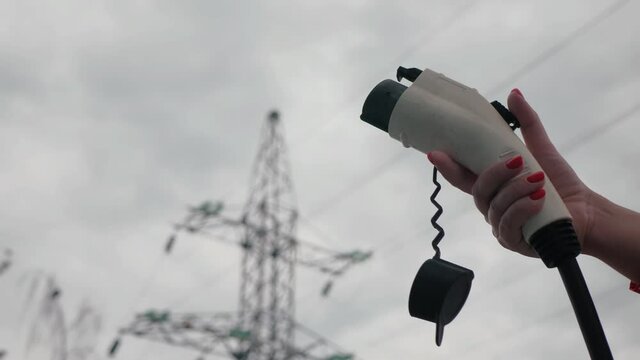 Female hands shows socket type 1, power supply cable to charge EV, close up. Electric car plug on blurred High voltage electric tower line on background. Eco-friendly alternative energy concept