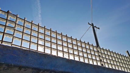Fototapeta premium Fluffy frost on a metal wire fence against the background of an electric pole and blue sky
