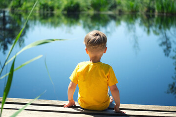 Lonely child boy sitting on wooden pier near lake on sunny summer day. Toddler kid in yellow...