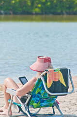 back view, medium distance of an adult female with a pink beach hat, sitting on a beach chair reading a bok on an electronic device , while taking  on a tropical, sandy beach