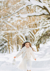 Fototapeta na wymiar Happy little girl runs on a forest path with a lantern in her hand. Christmas time. Sunny winter day.
