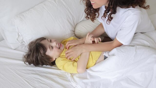 Cropped image of cheery mother cuddling and tickling little son lying in bed waking up in morning. Parent and child happy and lively game in bedroom. Carefree mummy and kid have fun at home concept