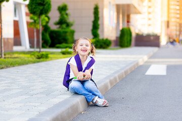 a happy child schoolgirl with a backpack and textbooks in her hands is sitting on the curb at the school, the concept is back to school