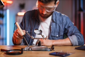 Focused bearded man in protective glasses sitting at table and soldering video card from modern...