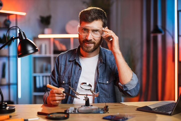 Caucasian young man in protective glasses using soldering iron and magnifying glass while repairing...