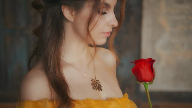 Girl beauty fantasy princess in yellow medieval silk dress holding flower red rose in hands. Background of old gothic stydio style of castle room. Fairy tale queen. Happy woman mystery smile on face.