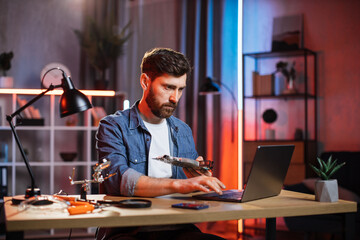 Obraz na płótnie Canvas Young bearded man with non working video card in hand searching in internet the new one. Caucasian guy in casual wear sitting at table and using modern laptop.