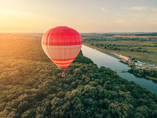 Hot air red balloon flies over the river and forest in the evening