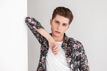Handsome portrait young man with serious face with clean skin with hairstyle in T-shirt in shirt fashionable vintage floral print near vintage white wall in room. Attractive fashion model guy indoors.