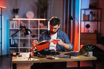 Focused bearded man in denim shirt sitting at table and repairing remote controlled car. Caucasian...