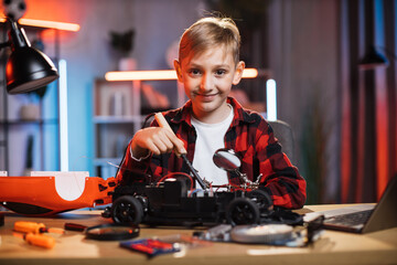Portrait of smiling little boy sitting at table and soldering remote controlled red car. Caucasian child in casual wear fixing favorite toy by himself.
