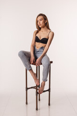 portrait of sexy asian woman with long hair posing in black lingerie, blue jeans on white studio background with bare feet. skinny pretty girl in bra sitting on chair. attractive female sits on stool