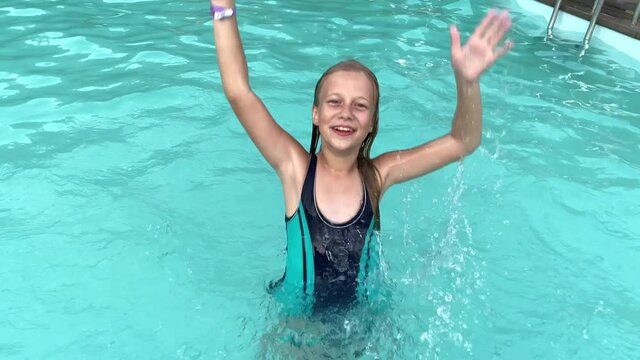 Cute blonde girl having fun in swimming pool. Child closing eyes and smiling while splashing water with her hands in slow motion