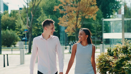 Multi-ethnic couple shyly looking at each other while walking the street, holding hands and talking. Relationship concept.