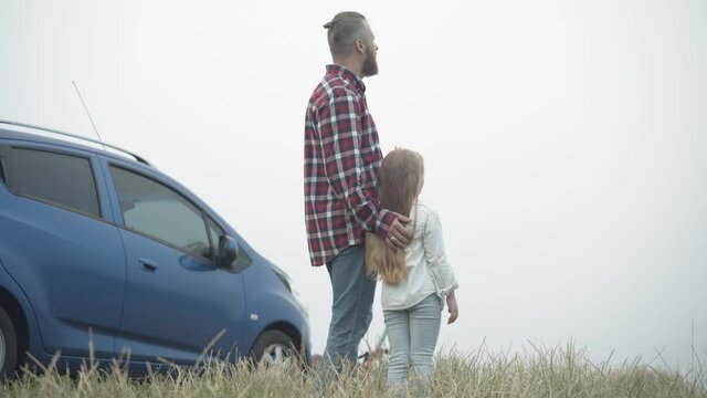 Side view of Caucasian bearded young father stroking hair of little daughter admiring beauty of misty landscape outdoors. Happy relaxed man and girl talking resting outdoors. Travelling and unity
