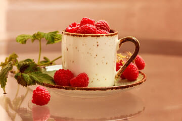 A cup full of fresh ripe red raspberries, green leaves  on a saucer in the background. Berry still life
