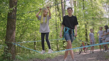 Small girl in climbing equipment in a rope Park. Group of Caucasian children training at boot camp. In the children camp, children are taught to overcome obstacles with the help of a rope crossing - Powered by Adobe