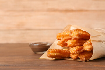 Tasty churros in parchment and melted chocolate sauce on wooden background, closeup
