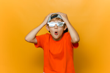Obraz na płótnie Canvas a child in a cap and glasses for watching movies grabbed his head with both hands and twisted his lips into a tube and shows emotions of surprise