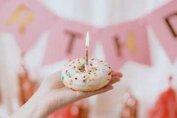 Delicious birthday donut with candle in hand on background of pink garland and decorations in...