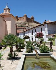 square in the town of La Calahorra with a fountain and plants next to the church with the castle of La Calahorra on a hill in the background. Some of the plants are small palm trees